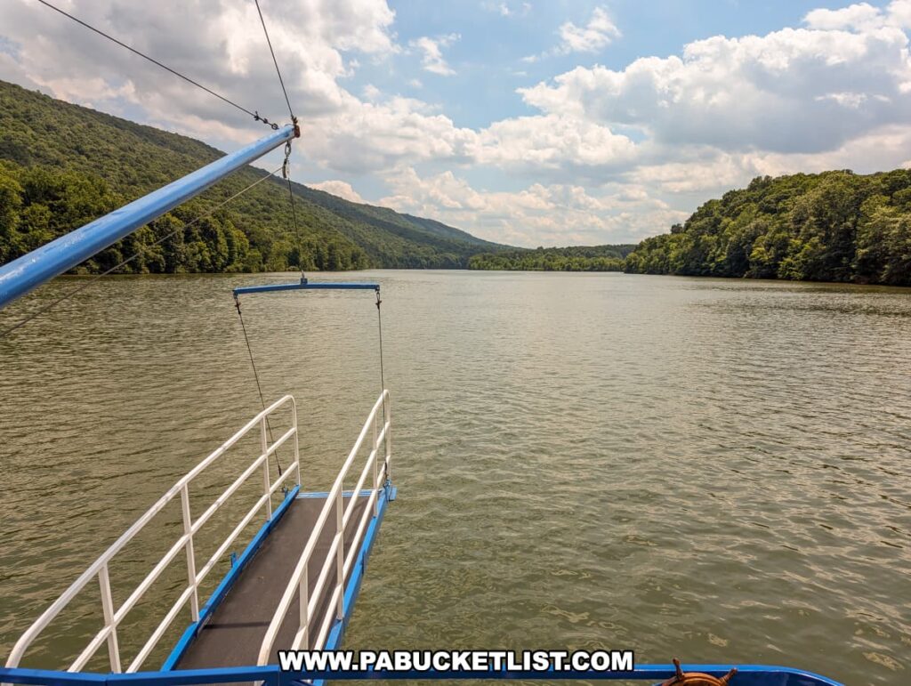 A view from the Proud Mary Showboat showing the boarding ramp extending over Raystown Lake. The calm waters are surrounded by lush, forested hills under a partly cloudy sky. The perspective highlights the peaceful and scenic nature of the lake.