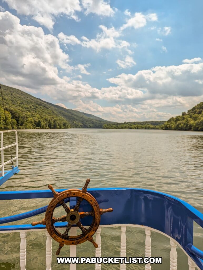 A view from the Proud Mary Showboat showing a decorative wooden ship's wheel mounted on the railing, with the boarding ramp extending over the calm waters of Raystown Lake. The lake is surrounded by lush, forested hills under a partly cloudy sky, creating a serene and picturesque atmosphere.
