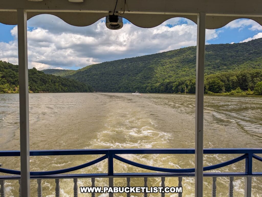 A view from the covered area at the stern of the Proud Mary Showboat, looking out over Raystown Lake. The boat's wake creates ripples in the water, with lush, green hills on either side under a partly cloudy sky. The railing and roof of the covered area frame the scenic landscape.