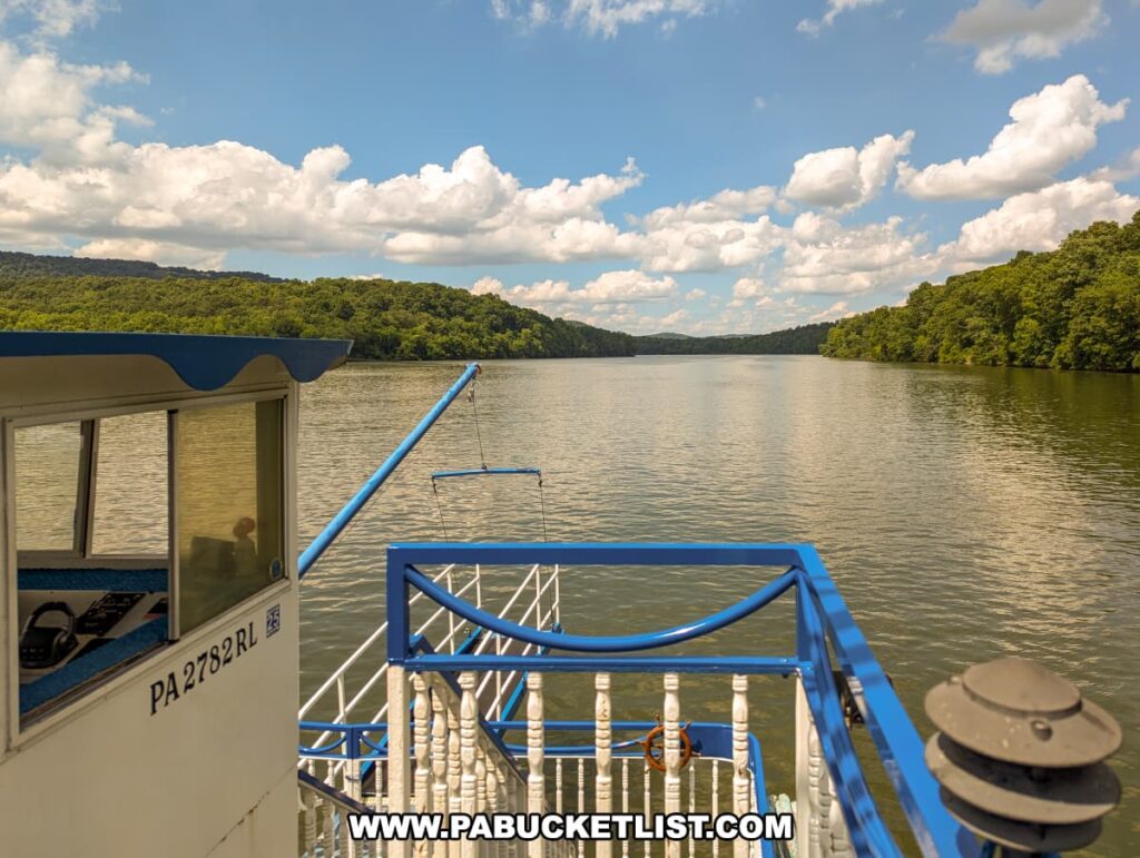 A view from the upper level of the Proud Mary Showboat, showing the boarding ramp and calm waters of Raystown Lake. The boat's railing frames the scene, with lush, green hills on either side under a bright blue sky with fluffy white clouds. The cabin and pilot house are visible on the left side of the image.
