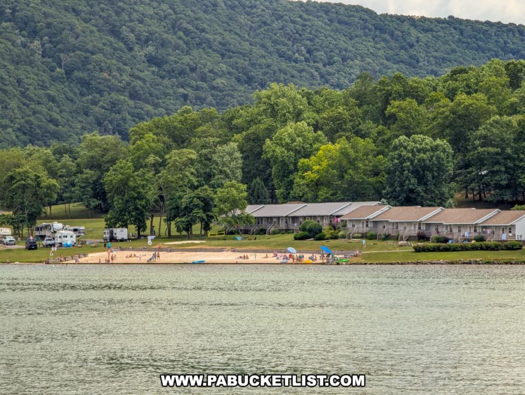 A view from the Proud Mary Showboat of a lakeside beach at Raystown Lake. The beach is populated with people enjoying the water and sand, surrounded by a lush green landscape. Nearby, there are several buildings and RVs, with forested hills providing a picturesque backdrop.