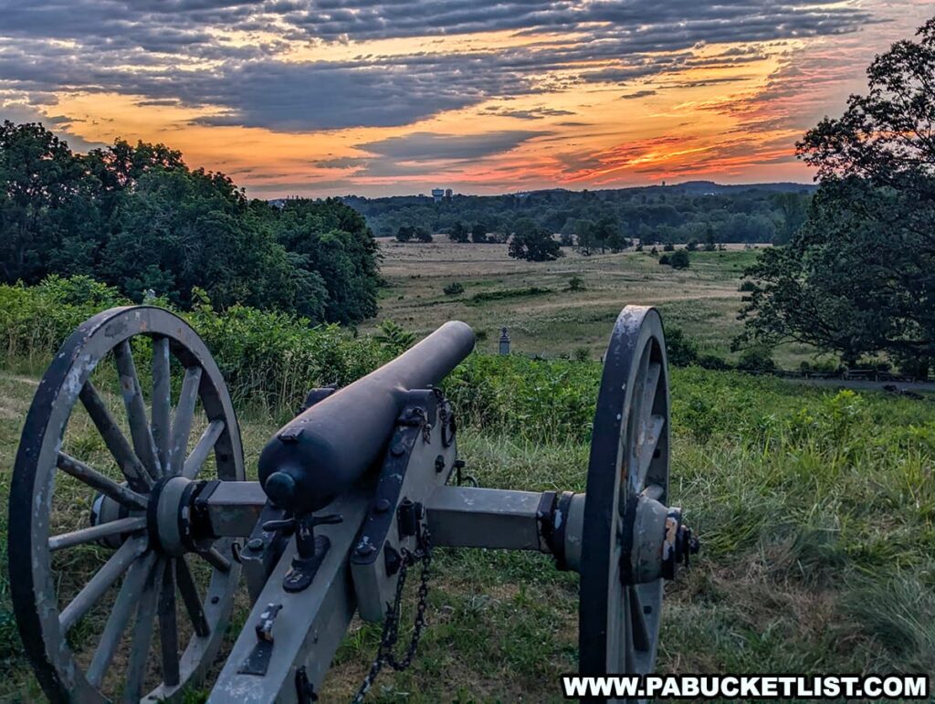 The photo captures a dramatic sunrise over East Cemetery Ridge in Gettysburg National Military Park. A historic cannon is prominently featured in the foreground, pointing towards the rolling fields and distant trees. The sky is ablaze with vibrant colors, ranging from deep oranges and reds near the horizon to softer blues and purples higher up, with clouds adding texture to the scene. The lush greenery and expansive landscape create a serene and reflective atmosphere, emphasizing the historical significance of this iconic battlefield.