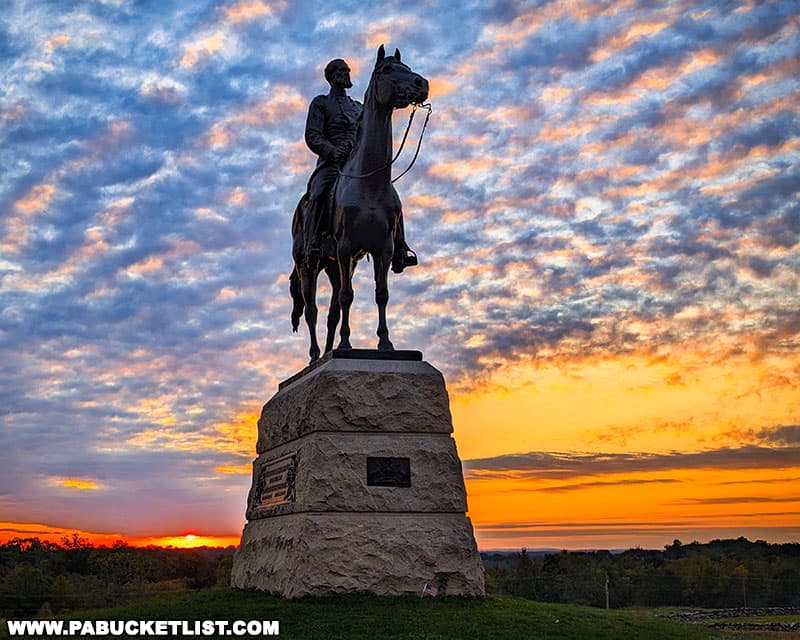 The photo showcases the equestrian statue of General George Meade at Gettysburg National Military Park during a stunning sunrise. The statue, depicting General Meade on horseback, stands atop a large stone pedestal with inscriptions. The sky behind the statue is filled with vibrant colors, ranging from deep oranges and yellows near the horizon to purples and blues higher up, with scattered clouds adding depth to the scene. The silhouette of the statue against the colorful dawn sky creates a powerful and dramatic image, emphasizing the historical significance and solemn beauty of this landmark.