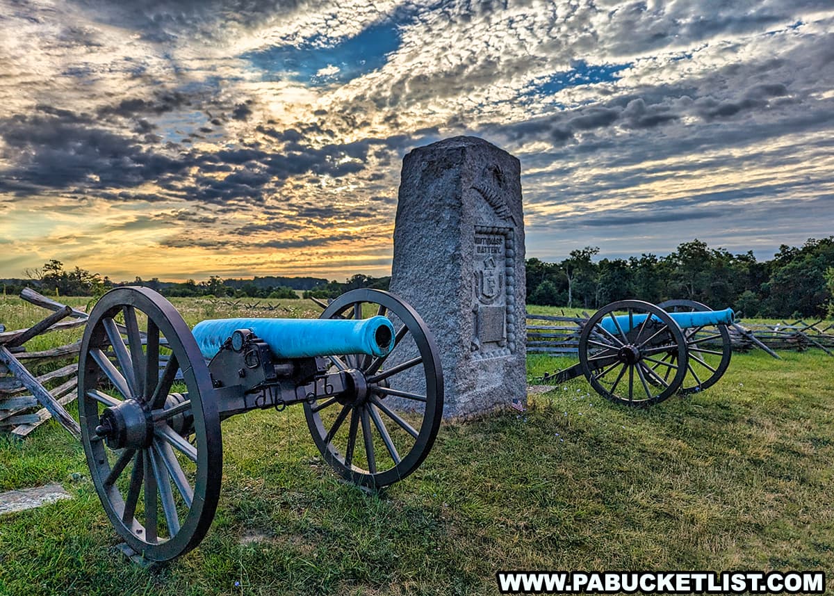 The photo captures a pair of historic cannons flanking a stone monument on the Gettysburg battlefield at sunrise. The cannons, with their blue-painted barrels, stand on lush green grass next to a split-rail fence. The monument features detailed engravings commemorating the artillery unit. The sky above is filled with dramatic clouds, painted with the warm colors of the rising sun, creating a stunning backdrop. The peaceful and reflective scene highlights the historical significance and natural beauty of this iconic location.