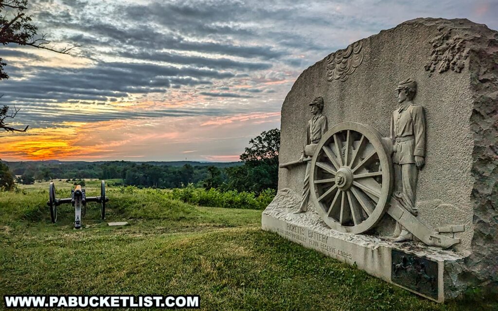 The photo captures the Ricketts Battery monument at East Cemetery Hill in Gettysburg National Military Park during sunrise. The monument features a detailed bas-relief sculpture of two soldiers standing beside a cannon wheel, carved into a large stone. In the background, a historic cannon points towards the expansive battlefield, framed by lush greenery and distant trees. The sky is painted with the warm hues of the rising sun, with dramatic clouds adding depth to the scene. The peaceful and reflective atmosphere underscores the historical significance of this location.