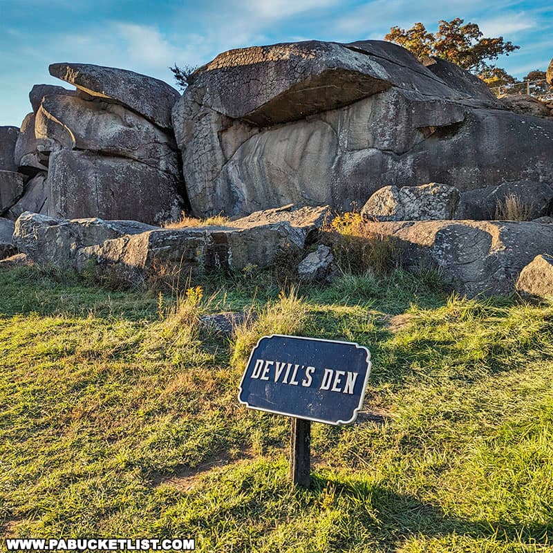 The photo shows the rocky formation known as Devil's Den in Gettysburg National Military Park. The large, weathered boulders create an imposing and rugged landscape. In the foreground, a sign labeled "Devil's Den" is placed on a grassy area, marking the historic site. The sunlight casts warm tones on the rocks and grass, highlighting the natural beauty and historical significance of this location on the battlefield. Trees and additional rock formations can be seen in the background under a clear blue sky.