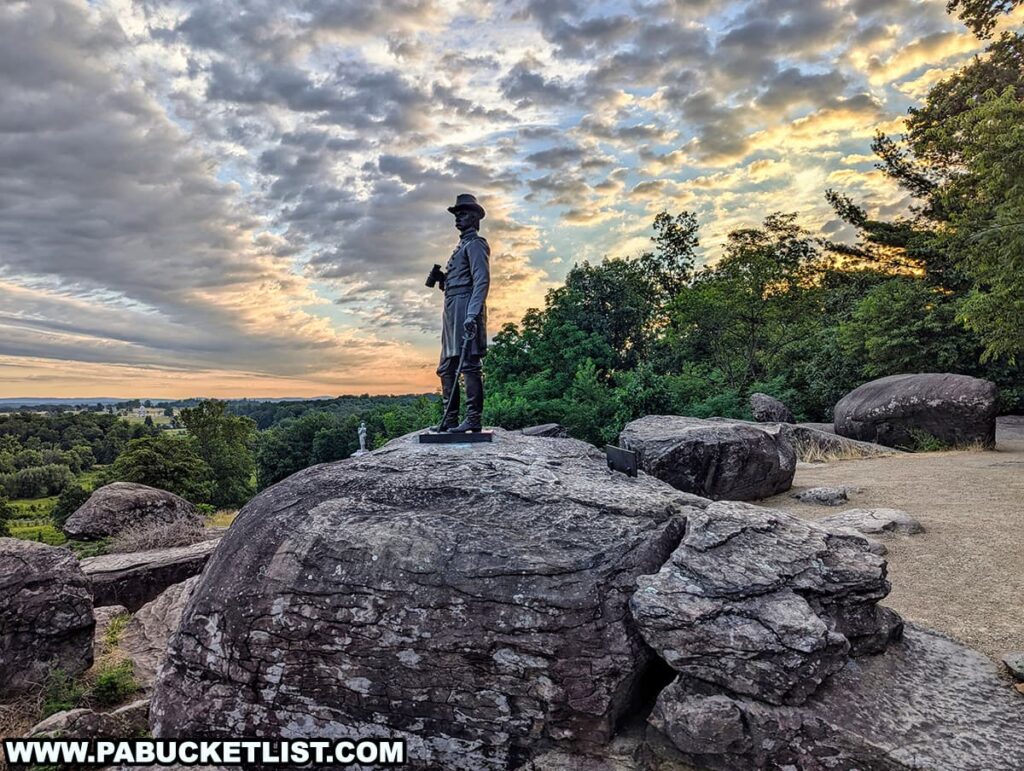 The photo captures the Warren Monument at sunrise on Little Round Top in Gettysburg National Military Park. The statue of General Gouverneur K. Warren stands atop a large rock, overlooking the battlefield. The statue shows Warren holding binoculars and gazing into the distance, symbolizing his role in the battle. Surrounding the monument are additional large boulders and lush green trees. The sky is filled with dramatic clouds illuminated by the rising sun, casting a warm glow over the landscape. The scene reflects the historical significance and natural beauty of this iconic location.