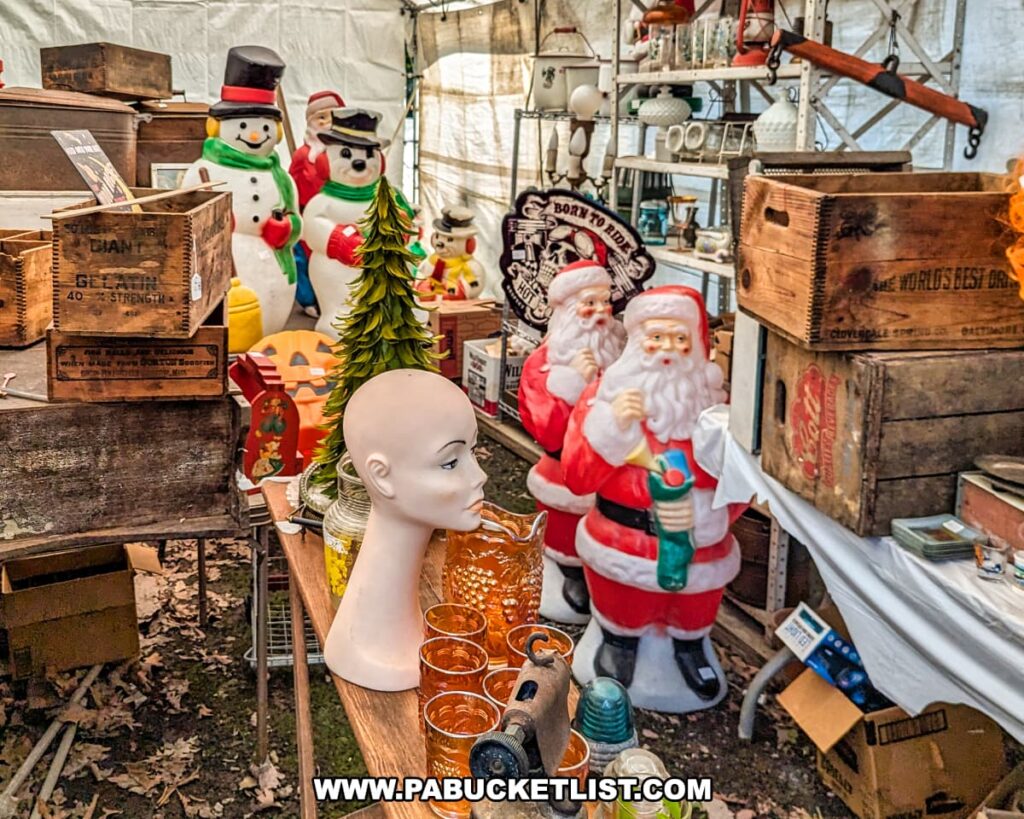 A vendor tent at Shupps Grove Flea Market in Lancaster County, Pennsylvania, is filled with a nostalgic collection of vintage holiday decorations and eclectic items. Prominently featured are several classic plastic Santa Claus and snowman figures, surrounded by old wooden crates, glassware, and a mannequin head. The scene is a mix of Christmas cheer and vintage charm, with a variety of unique items displayed on shelves and tables. The tent is part of the market's wooded, park-like setting, which adds to the cozy and inviting atmosphere of this long-standing flea market, operational since 1962.