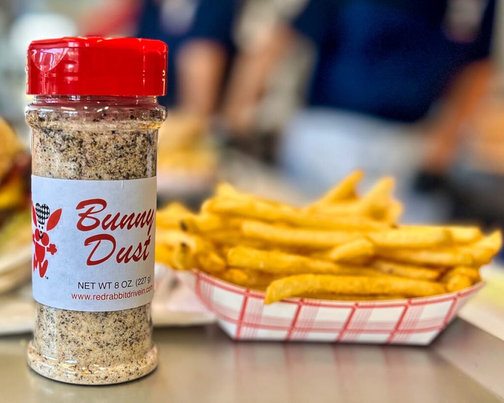 A close-up photograph of a bottle of Bunny Dust seasoning in the foreground at the Red Rabbit Drive-In Restaurant in Duncannon, PA. The seasoning bottle has a red cap and a label featuring the restaurant’s signature bunny mascot, with bold red lettering. In the background, a serving of crispy golden French fries, generously coated with Bunny Dust, sits in a red-and-white checkered paper tray. The blurred background captures the bustling energy of the restaurant, hinting at the classic 1950s-style drive-in atmosphere that Red Rabbit is known for.