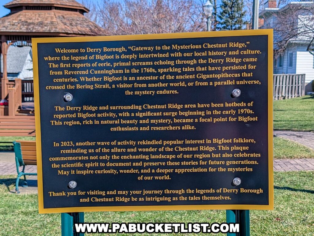 A photograph of an informational plaque next to the Derry Bigfoot statue in Derry Borough, Westmoreland County, Pennsylvania. The plaque, mounted on a metal stand with a gold border, provides historical context about Bigfoot lore in the region, referring to Derry Borough as the "Gateway to the Mysterious Chestnut Ridge." It details early reports of eerie, primal screams from the 1760s, the area's significance as a hotspot for Bigfoot sightings since the 1970s, and a resurgence of interest in 2023. The text discusses theories about Bigfoot’s origins and highlights the region's natural beauty and mystery. The background features a wooden gazebo, park benches, lampposts, and residential houses under a clear blue sky, emphasizing the plaque's location in a public park setting.