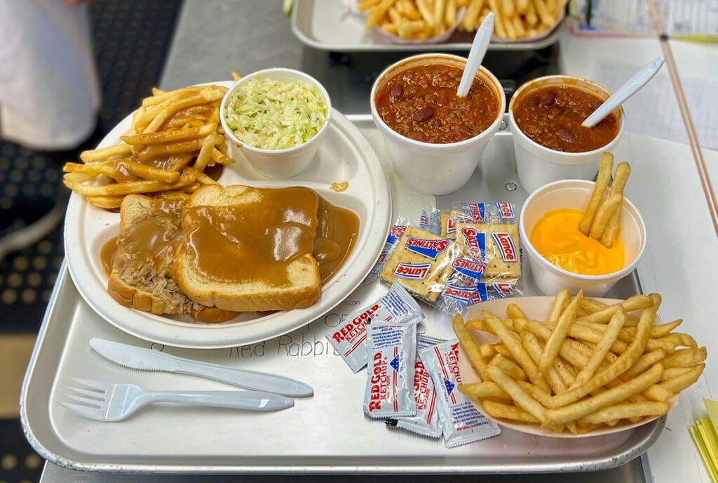 A tray of food from the Red Rabbit Drive-In Restaurant in Duncannon, PA, featuring a hearty meal with classic diner-style comfort foods. The main plate includes an open-faced roast beef sandwich on white bread, generously smothered in rich brown gravy, accompanied by a side of crispy French fries also topped with gravy and a cup of fresh coleslaw. Additional items on the tray include two cups of thick, meaty chili with kidney beans, a serving of golden fries with a cup of creamy nacho cheese sauce, and individually wrapped saltine crackers. Several packets of Red Gold ketchup and plastic utensils are also provided. In the background, another tray of food with more French fries is visible, adding to the nostalgic and welcoming feel of this 1950s-style drive-in restaurant.