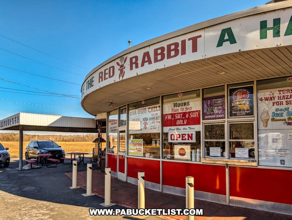 The exterior of the Red Rabbit Drive-In Restaurant in Duncannon, PA, showcasing its classic 1950s-style design with a curved overhang, red and white color scheme, and large front windows filled with menu signs and promotional posters. The restaurant's iconic bunny mascot is displayed on the signage above the entrance. A covered carhop service area is visible to the left, with picnic tables available for outdoor dining. A few vehicles are parked nearby, highlighting the retro drive-in experience. The bright blue sky and open fields in the background emphasize the nostalgic roadside charm of this beloved Pennsylvania eatery.