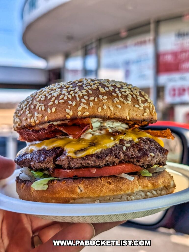 A close-up of a freshly made Bunny Burger from the Red Rabbit Drive-In Restaurant in Duncannon, PA, held on a white paper plate with the restaurant’s exterior visible in the background. The burger features two juicy beef patties topped with melted cheddar cheese, crispy bacon, fresh lettuce, a thick slice of tomato, and a dollop of special sauce, all sandwiched between a toasted sesame seed bun. The vibrant colors and textures of the ingredients highlight the classic, made-to-order quality of this signature menu item at the nostalgic 1950s-style drive-in restaurant.