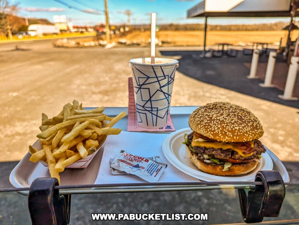 A classic carhop tray meal at the Red Rabbit Drive-In Restaurant in Duncannon, PA, featuring a Bunny Burger, a side of crispy French fries, and a chocolate milkshake. The Bunny Burger, served on a sesame seed bun, is topped with melted cheddar cheese, crispy bacon, lettuce, tomato, and special sauce. The golden fries are presented in a red-and-white checkered paper tray, accompanied by a packet of Red Gold ketchup. The meal is set on a nostalgic drive-in tray attached to a car window, with the restaurant’s outdoor seating area and rural Pennsylvania landscape visible in the background, capturing the timeless charm of this 1950s-style roadside eatery.