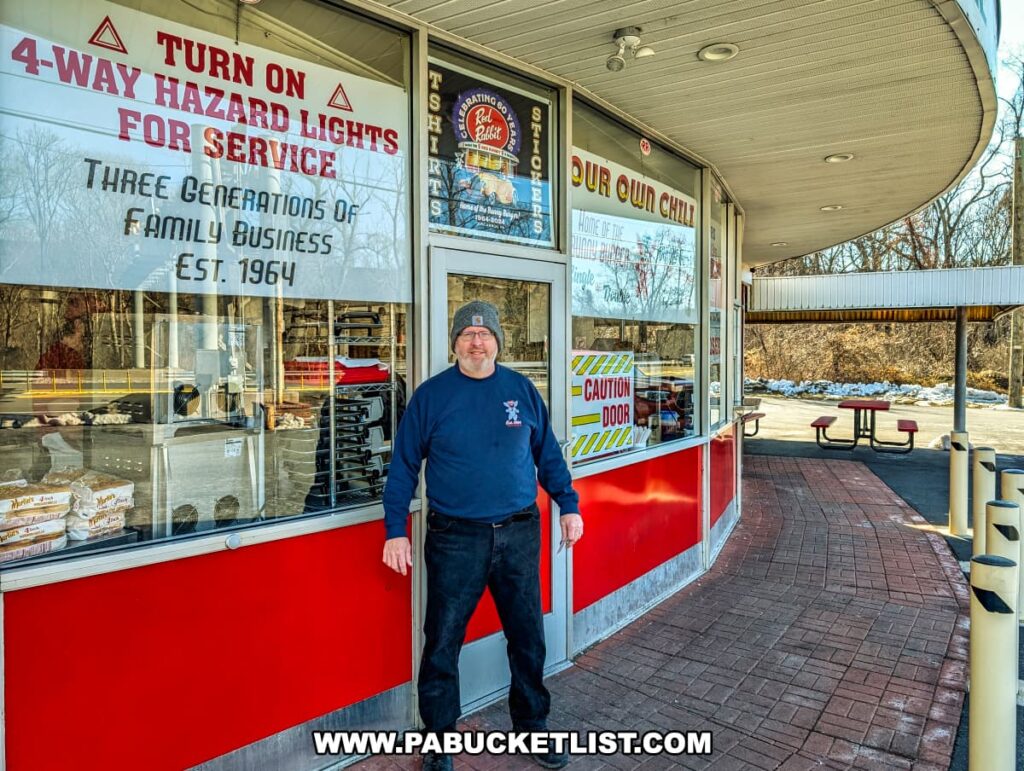 A man in a navy blue sweatshirt and gray beanie stands in front of the Red Rabbit Drive-In Restaurant in Duncannon, PA, smiling at the camera. Behind him, the restaurant’s curved overhang and red-and-white exterior showcase its classic 1950s-style architecture. The large front windows display signage advertising the restaurant’s signature Bunny Burger, house-made chili, and Bunny Dust seasoning. A sign highlights the establishment’s history as a three-generation family business since 1964, and another instructs customers to turn on their four-way hazard lights for carhop service. The outdoor seating area with picnic tables and a covered drive-in service section is visible in the background, adding to the nostalgic charm of this beloved Pennsylvania roadside eatery.