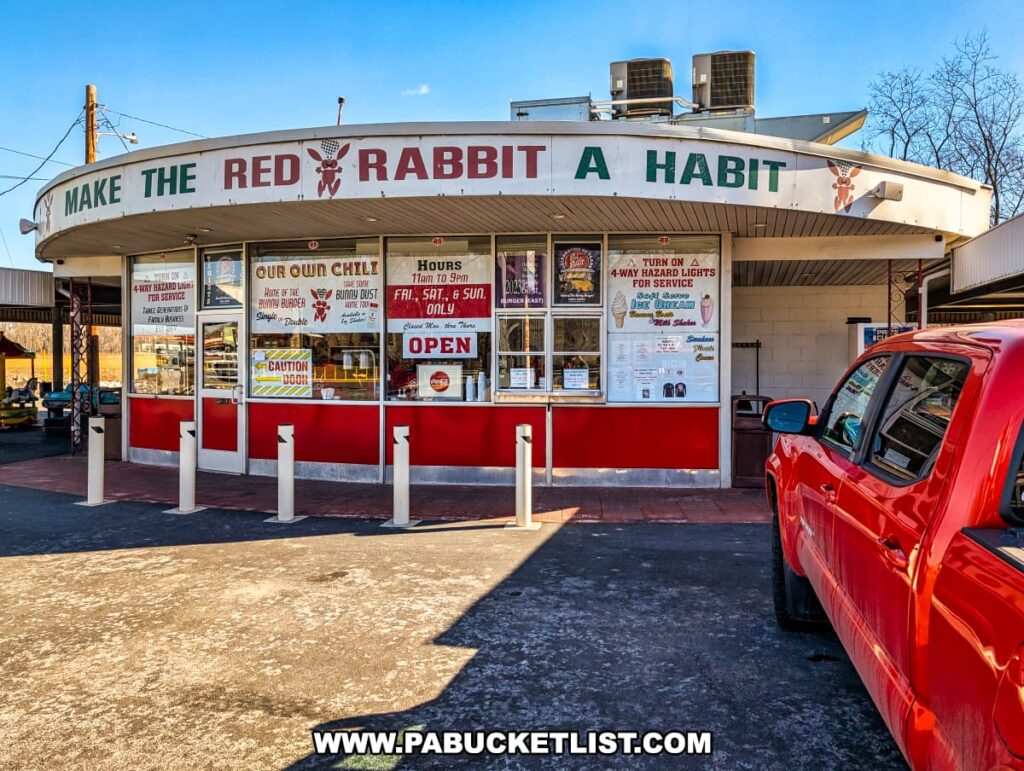 The front view of the Red Rabbit Drive-In Restaurant in Duncannon, PA, showcasing its retro 1950s-style architecture with a curved overhang displaying the slogan "Make the Red Rabbit a Habit." The exterior features red and white paneling with large windows covered in menu signs, highlighting signature items such as the Bunny Burger, Bunny Dust, and homemade chili. A red pickup truck is parked in front, while the carhop service area and outdoor picnic tables are visible in the background. The clear blue sky and rural setting add to the nostalgic charm of this classic Pennsylvania roadside eatery.