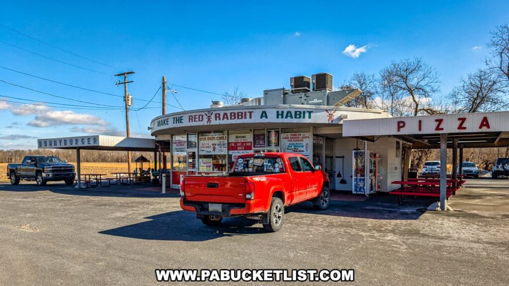 A wide-angle view of the Red Rabbit Drive-In Restaurant in Duncannon, PA, capturing its nostalgic 1950s-style design. The curved overhang displays the slogan "Make the Red Rabbit a Habit," while large front windows showcase menu items like the famous Bunny Burger and house-made chili. A red pickup truck is parked in front, adding to the classic roadside diner atmosphere. To the left, a covered carhop service area with picnic tables invites outdoor dining, while a "Pizza" sign marks another section of the restaurant. The bright blue sky and rural Pennsylvania backdrop emphasize the charm of this beloved family-owned drive-in eatery.