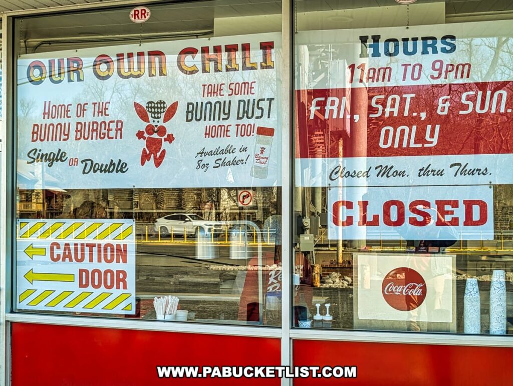 A close-up of the front window at the Red Rabbit Drive-In Restaurant in Duncannon, PA, displaying colorful signage promoting the restaurant’s signature offerings. The signs advertise "Our Own Chili," the famous "Bunny Burger" available as a single or double, and "Bunny Dust" seasoning available in an 8-ounce shaker to take home. Another sign indicates the restaurant's hours, stating it is open only on Fridays, Saturdays, and Sundays from 11 AM to 9 PM and closed Monday through Thursday. A bold "Caution Door" warning with yellow arrows is also visible. Reflections of cars passing by on the road can be seen in the glass, adding to the nostalgic roadside charm of this classic 1950s-style drive-in.