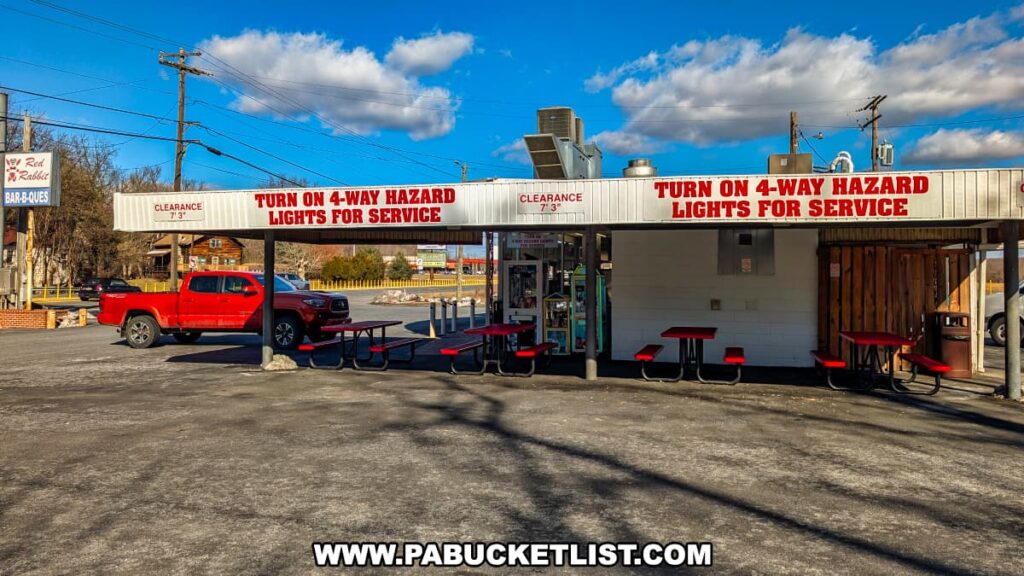 A side view of the Red Rabbit Drive-In Restaurant in Duncannon, PA, highlighting the classic carhop service area. The covered drive-in section features bold red lettering instructing customers to "Turn on 4-way hazard lights for service," a signature feature of the restaurant's retro dining experience. Several red picnic tables are set up for outdoor seating, while a red pickup truck is parked nearby. The Red Rabbit’s roadside sign is visible in the background along with Route 322, emphasizing the nostalgic charm of this beloved 1950s-style eatery. The bright blue sky and scattered clouds add to the inviting atmosphere of this Pennsylvania roadside landmark.