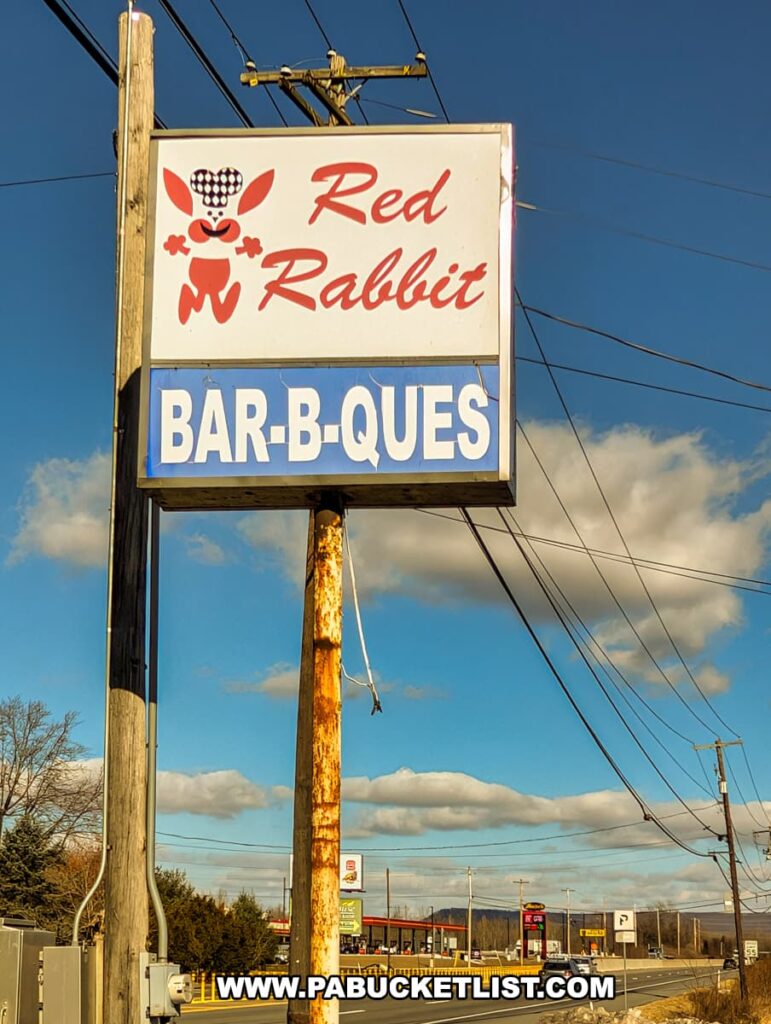 A roadside sign for the Red Rabbit Drive-In Restaurant in Duncannon, PA, standing tall against a bright blue sky with scattered clouds. The sign features the restaurant’s iconic red bunny mascot wearing a checkered hat, along with bold red lettering spelling out “Red Rabbit.” Below, a blue panel highlights one of the restaurant’s specialties, “BAR-B-QUES.” The sign is mounted on a weathered utility pole, with power lines stretching overhead. In the background, Route 322 runs past the restaurant, with a few businesses and gas stations visible in the distance, emphasizing the nostalgic roadside charm of this classic 1950s-style drive-in eatery.