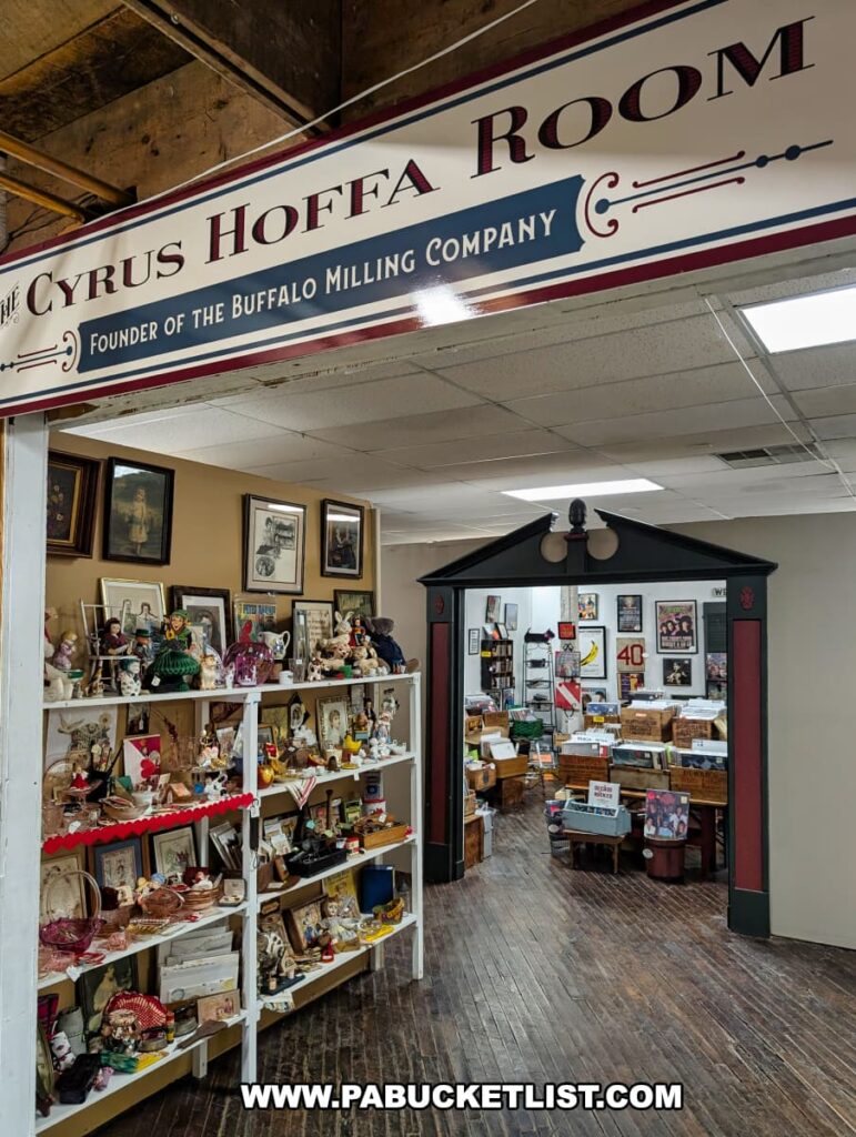 The entrance to the Cyrus Hoffa Room inside Rollermills Antique Marketplace in Lewisburg, PA, named after the founder of the Buffalo Milling Company, featuring shelves filled with antique figurines, framed artwork, and collectibles, leading into a larger room with vintage memorabilia, books, and historical artifacts displayed on wooden floors within the restored 1880s flour mill.
