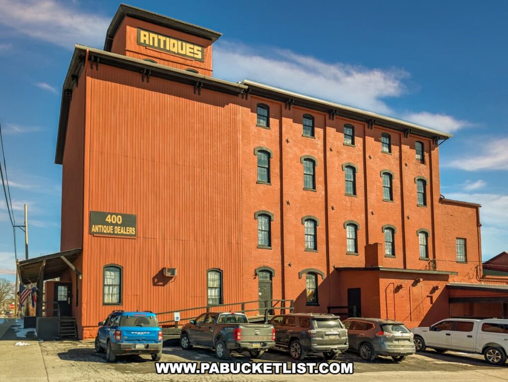The exterior of Rollermills Antique Marketplace in Lewisburg, PA, a large multi-story red brick building that was once an 1880s flour mill, now home to 400 antique dealers, with signage advertising antiques and a parking area in front under a clear blue sky.