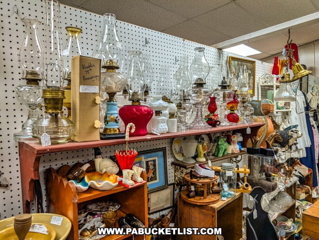 A vendor booth inside Rollermills Antique Marketplace in Lewisburg, PA, displaying a collection of vintage oil lamps, glassware, and decorative antiques, with shelves filled with unique collectibles, ceramic dishes, and rustic home decor, all arranged against a pegboard backdrop in the historic 1880s flour mill.