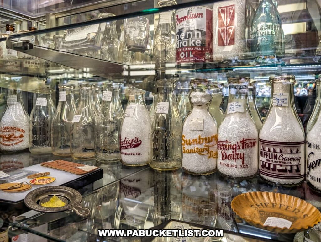 A glass display case inside Rollermills Antique Marketplace in Lewisburg, PA, featuring a collection of vintage milk bottles from regional dairies, embossed and painted with classic logos, alongside antique glassware, tins, and collectible memorabilia, showcasing the marketplace’s variety of historical items.