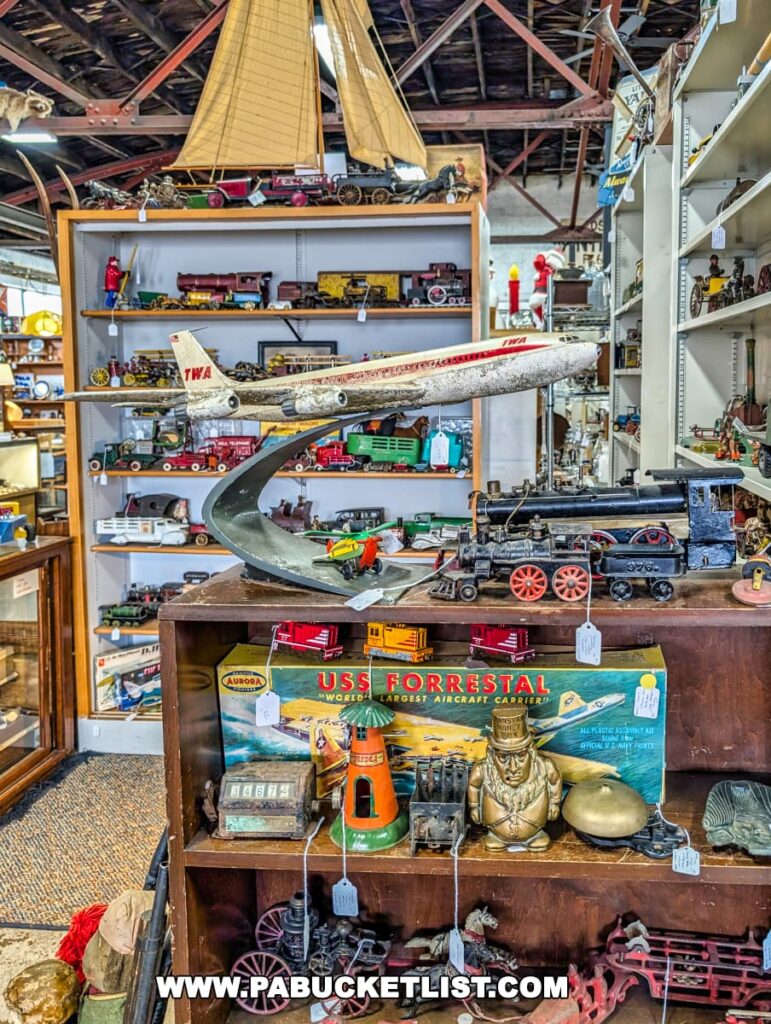 Display of vintage model toys and collectibles at the Duncansville Antiques Co-Op in Blair County, Pennsylvania, featuring a TWA airplane model on a stand, an Aurora USS Forrestal aircraft carrier model kit, antique trains, toy cannons, and various miniature vehicles arranged on wooden shelves.