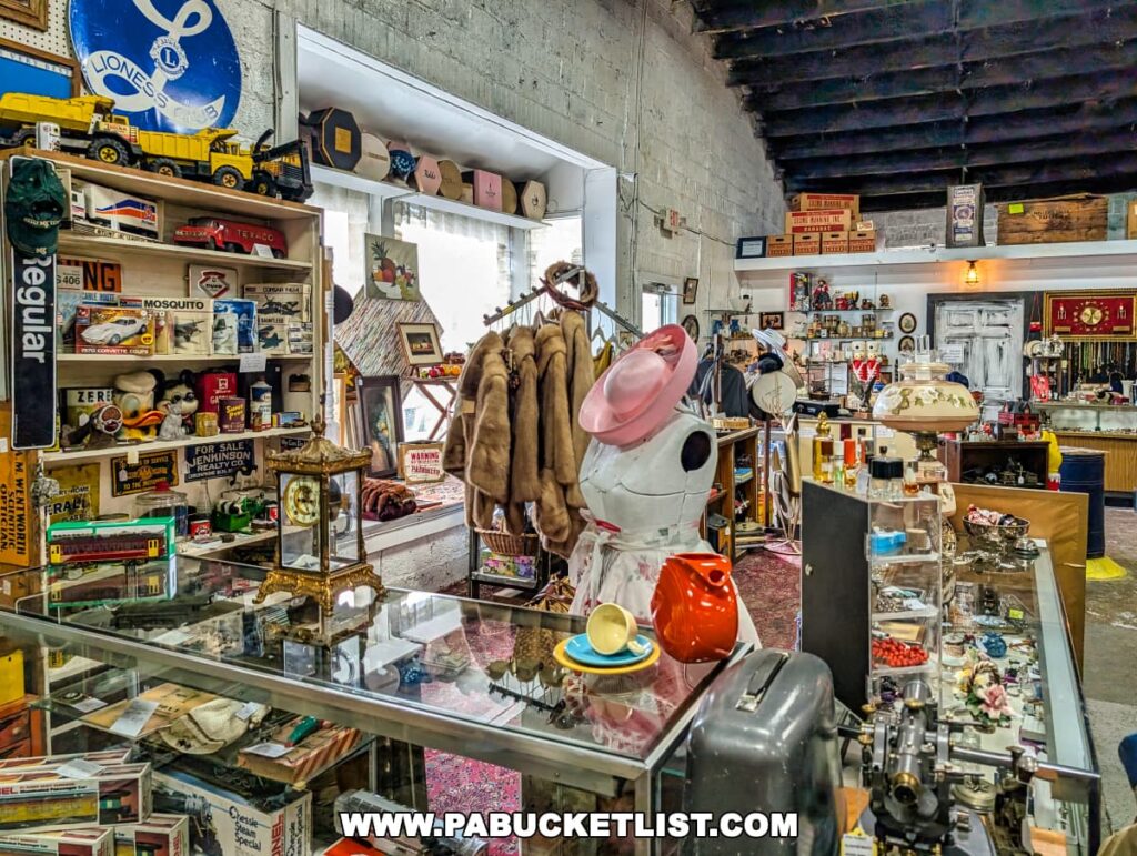 Interior of the Duncansville Antiques Co-Op in Blair County, Pennsylvania, featuring a diverse assortment of vintage collectibles, including a mannequin dressed in a retro dress and pink hat, a rack of fur coats, glass display cases filled with jewelry and small antiques, and shelves stocked with old toys, advertising signs, and decorative items.