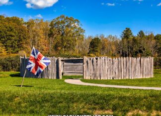 A reconstructed wooden stockade at Fort Necessity National Battlefield in Fayette County, Pennsylvania, stands against a backdrop of autumn foliage and a bright blue sky. A British flag with the red cross of St. George and the white saltire of St. Andrew flutters in the foreground, marking the site of the historic 1754 battle between British and French forces during the French and Indian War. The grassy landscape and winding pathway lead visitors to the entrance of the fort, evoking the setting where a young George Washington commanded his troops in a pivotal early conflict.