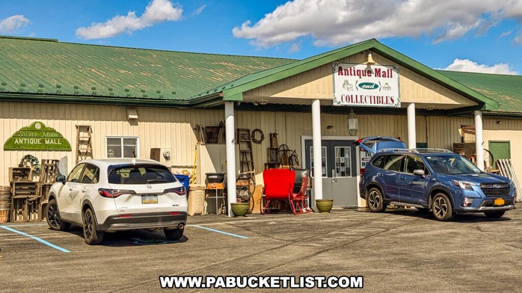 Exterior view of the Silver Moon Antique Mall, part of the Lewisburg Antique Malls in Union County, Pennsylvania, featuring a beige building with a green roof, antique items displayed near the entrance, a welcoming sign indicating "Antique Mall and Collectibles," and cars parked in front on a sunny day.