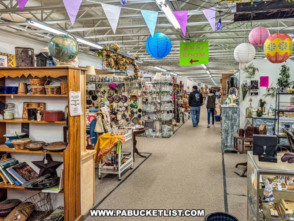 Interior scene at the Lewisburg Antique Malls in Union County, Pennsylvania, highlighting vendor booths filled with antiques and collectibles, including pottery, vintage kitchenware, glassware, books, decorative items, colorful lanterns, and festive banners, with shoppers exploring the aisles.