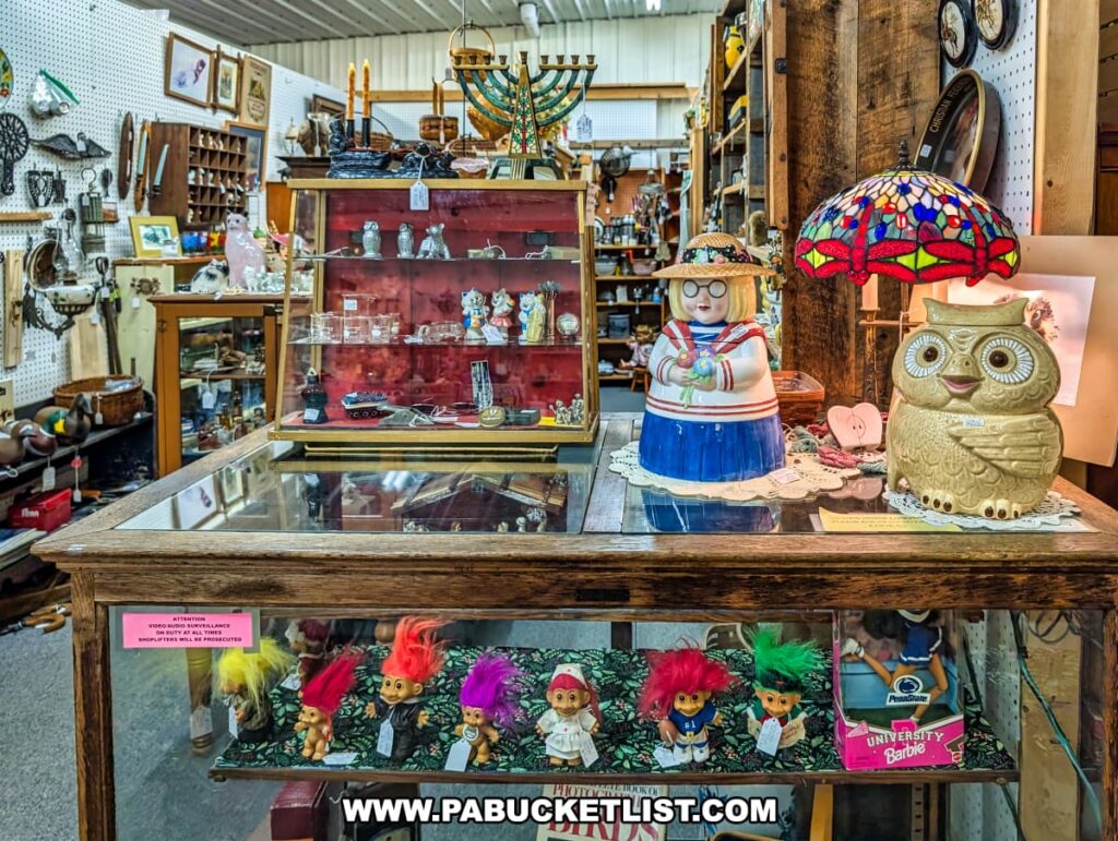 Vendor booth at the Lewisburg Antique Malls in Union County, Pennsylvania, displaying vintage collectibles including colorful troll dolls, character-themed cookie jars, decorative lamps, glassware, pottery, and other nostalgic items arranged neatly on glass display cases and shelves.