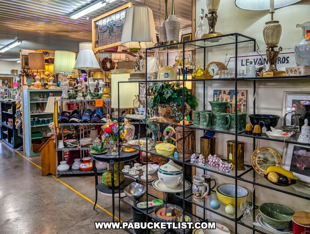 Vendor booth at the Lewisburg Antique Malls in Union County, Pennsylvania, showcasing a wide selection of vintage lamps, decorative glassware, ceramics, dishes, mugs, collectibles, and home décor items carefully arranged on shelves and racks along a well-lit aisle.