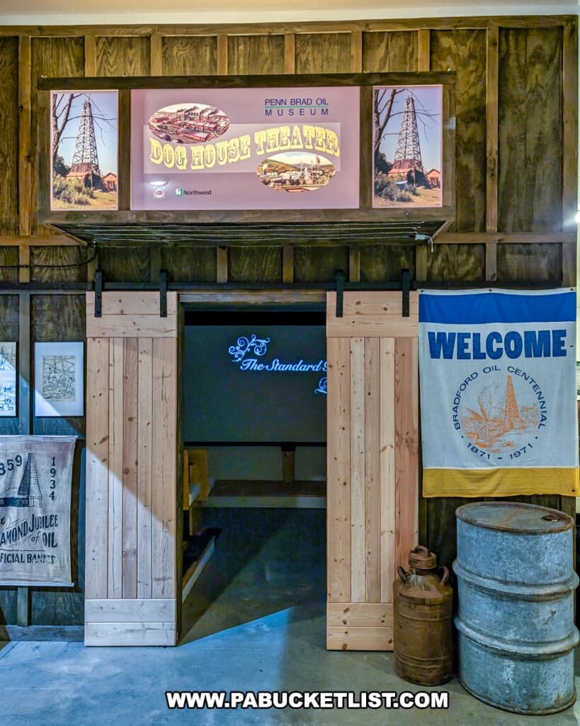 Entrance to the Dog House Theater inside the Penn Brad Oil Museum in McKean County, Pennsylvania, featuring sliding wooden barn doors leading to a small theater where visitors can watch a film about the history of the Bradford Oil Field; the scene also includes vintage oil industry artifacts such as an oil barrel, an old oil can, framed prints, and a banner commemorating the Bradford Oil Centennial from 1871 to 1971.