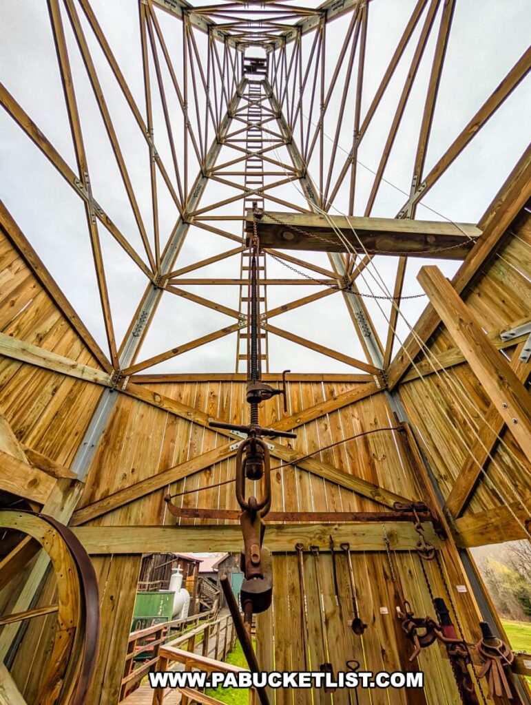 View looking upward from the base of a reconstructed wooden oil derrick at the Penn Brad Oil Museum in McKean County, Pennsylvania, showcasing the framework of the towering structure, pulleys, cables, and drilling equipment used in historic oil field operations, with various tools hanging on the wooden interior walls and a glimpse of additional outdoor exhibits visible in the background.