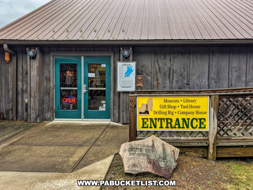 Exterior view of the entrance to the Penn Brad Oil Museum in McKean County, Pennsylvania, featuring a rustic wooden building with teal double doors, an illuminated open sign, and a large yellow sign directing visitors to the museum, library, gift shop, tool house, drilling rig, and company house; a commemorative stone marker for the Custer City Centennial from 1877 to 1977 is positioned in front of the entrance walkway.