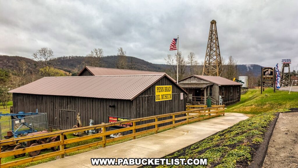 Exterior view of the Penn Brad Oil Museum in McKean County, Pennsylvania, featuring rustic wooden buildings with metal roofs, a large yellow sign identifying the museum, an American flag flying on a pole, and a tall wooden oil derrick representing the region's rich oil heritage; a sidewalk and wooden fence guide visitors to the entrance, while vintage oil field equipment is displayed outdoors against a backdrop of rolling hills and cloudy skies.