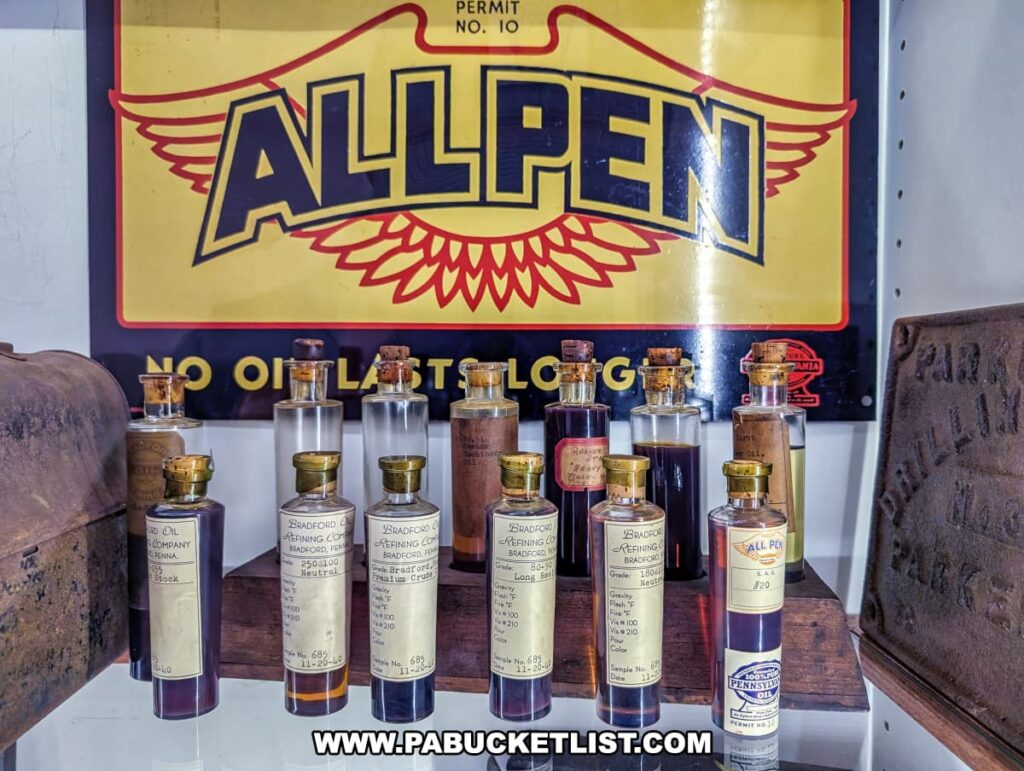 Display of vintage oil samples at the Penn Brad Oil Museum in McKean County, Pennsylvania, featuring glass bottles filled with various grades of crude oil and refined oil products from the Bradford Refining Company, each labeled with detailed information; the bottles are arranged in front of a colorful All Pen Motor Oil sign promoting Pennsylvania-grade oil with the slogan "No Oil Lasts Longer," highlighting the area's rich oil refining history.