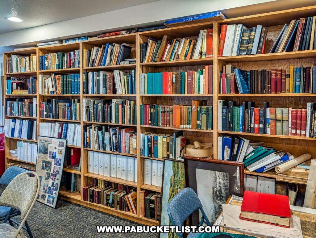 Research library inside the Penn Brad Oil Museum in McKean County, Pennsylvania, featuring tall wooden bookshelves filled with books, binders, photographs, and archival materials related to the history of the oil industry and the Bradford Oil Field; the room includes chairs for visitors, framed historical images, and displays of research resources documenting Pennsylvania's oil heritage.