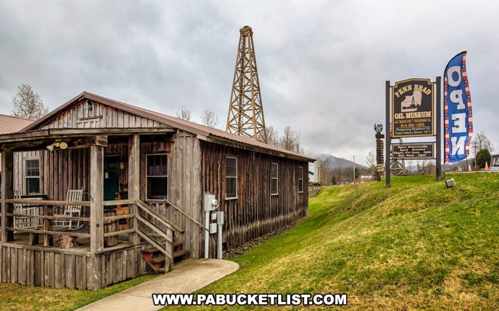Street view of the Penn Brad Oil Museum in McKean County, Pennsylvania, featuring a rustic wooden building with a covered front porch and rocking chairs, a tall wooden oil derrick in the background, and a large roadside sign identifying the museum and its seasonal hours of operation; an American flag-themed "OPEN" banner flutters nearby, and the scene is set against a backdrop of rolling hills and a cloudy sky.
