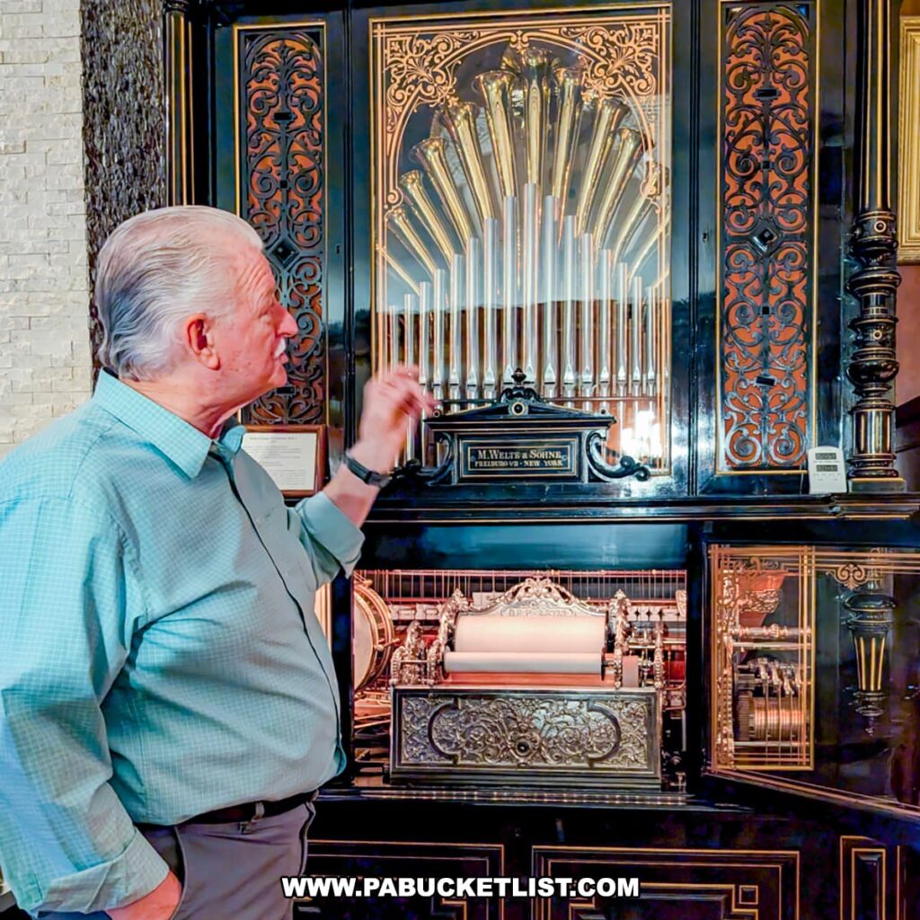A tour guide at the Bayernhof Museum gestures toward an ornate antique mechanical music machine with polished brass pipes, intricate woodwork, and a visible paper roll mechanism, highlighting the museum’s extensive collection of automatic musical instruments housed within the lavish mansion built by Charles B. Brown III.
