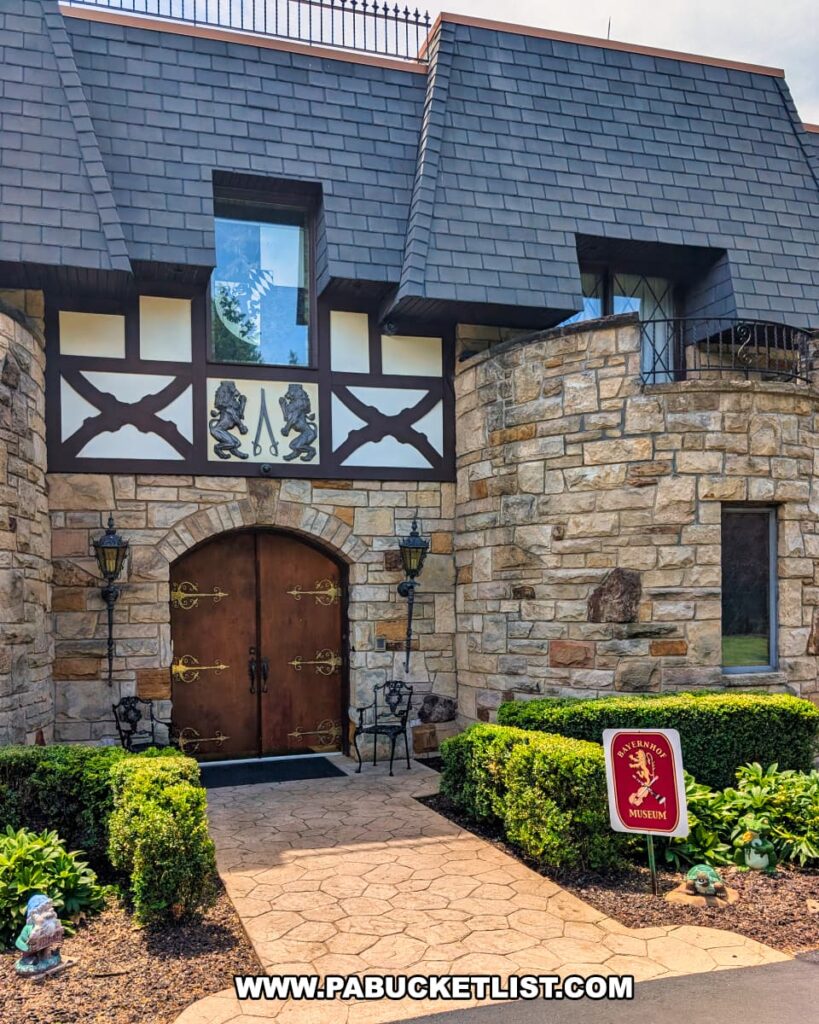 The front entrance of the Bayernhof Museum features a medieval-inspired arched wooden doorway with ornate brass hinges, flanked by stone walls and lanterns, with a manicured garden path leading to the door, Tudor-style detailing above, and a red museum sign displaying a lion crest welcoming visitors to this Bavarian-style mansion overlooking the Allegheny River.