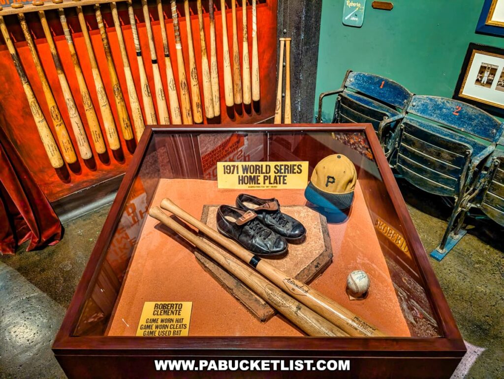 Display case at the Roberto Clemente Museum in Pittsburgh, PA, containing the 1971 World Series home plate along with Roberto Clemente’s game-worn cleats, game-used bats, and Pirates cap, surrounded by vintage stadium seats and a wall of wooden baseball bats, commemorating his MVP performance in the series.