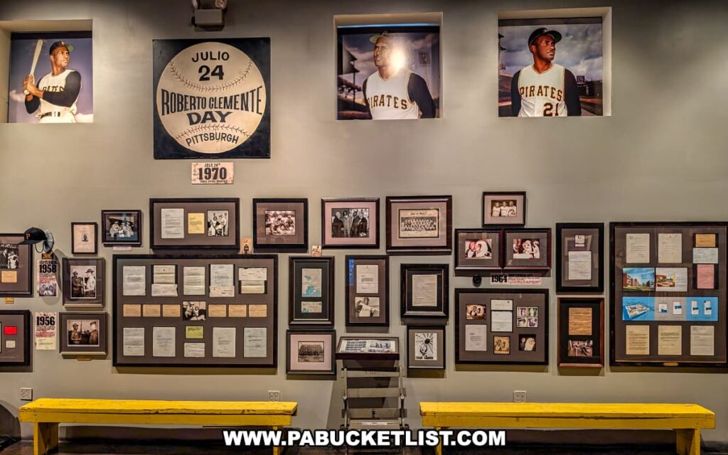 Wall display at the Roberto Clemente Museum in Pittsburgh, PA, showcasing framed photographs, personal letters, documents, and memorabilia beneath a large sign reading “Roberto Clemente Day – Julio 24,” with portraits of Clemente in his Pirates uniform displayed in recessed panels above bright yellow benches.