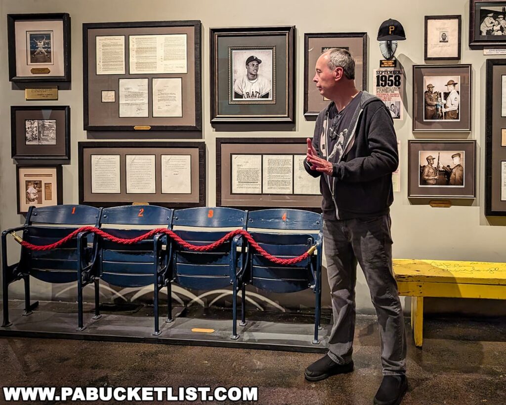 Docent at the Roberto Clemente Museum in Pittsburgh, PA, standing in front of a wall of framed documents and photographs related to Clemente’s life and career, next to a roped-off row of vintage stadium seats and a signed yellow bench beneath a Pittsburgh Pirates cap and memorabilia display.