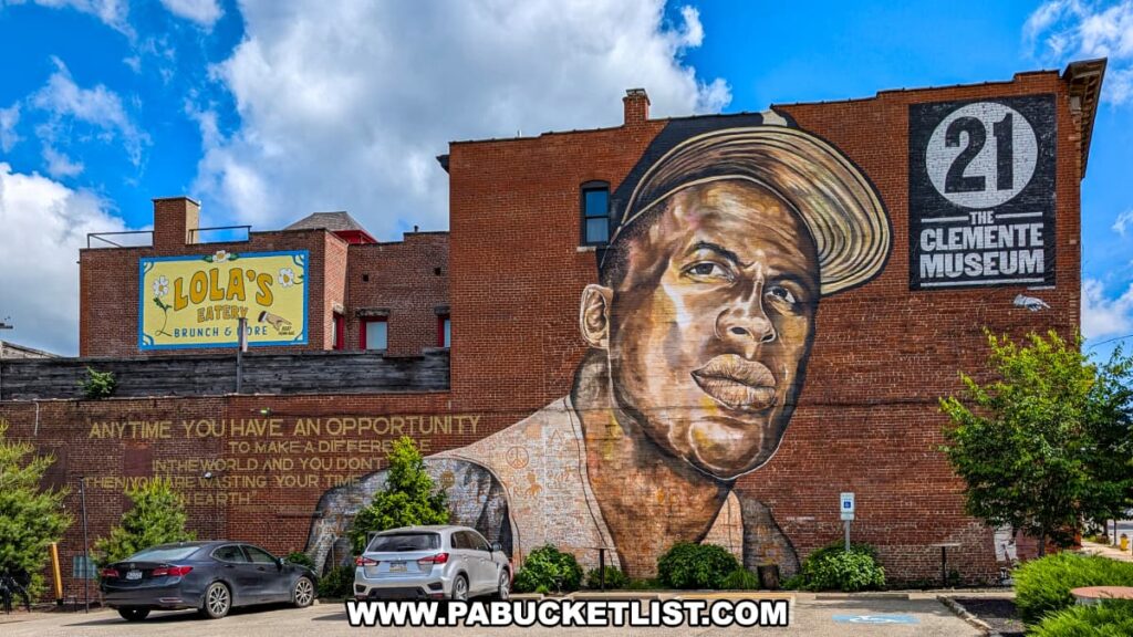 Large mural of Roberto Clemente on the exterior brick wall of the Roberto Clemente Museum in Pittsburgh, PA, with his number 21 and museum logo painted above, and an inspiring quote about making a difference in the world displayed alongside parked cars and nearby trees.