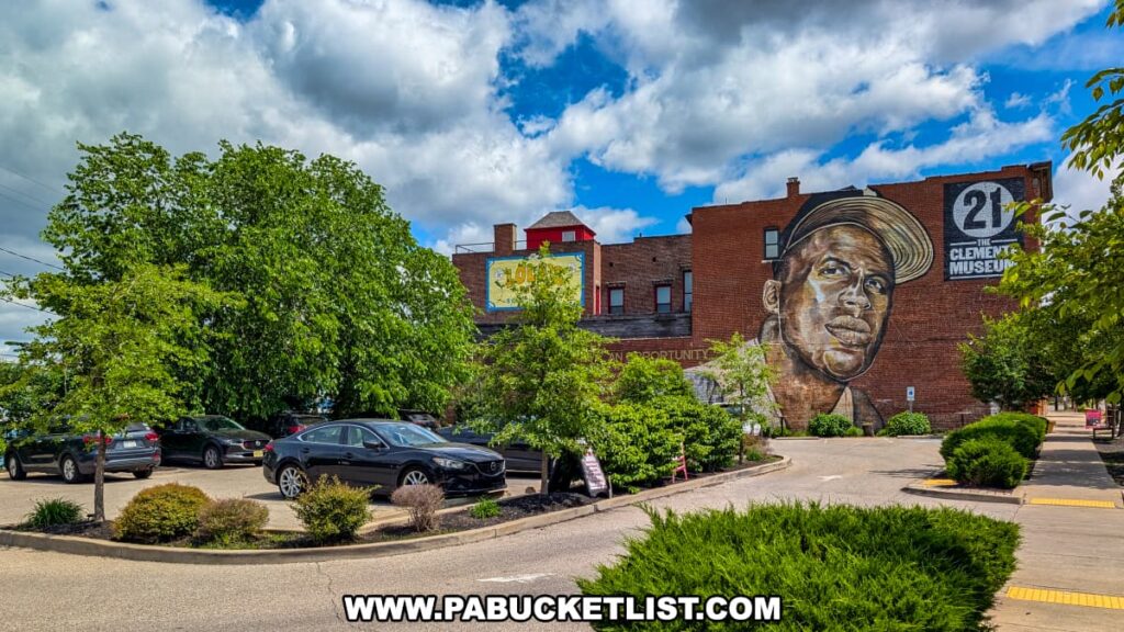 View of the Roberto Clemente Museum parking lot in Pittsburgh, PA, featuring a large mural of Clemente's face on the side of a brick building, with the number 21 and museum logo above, surrounded by lush trees, landscaped greenery, and parked cars under a partly cloudy sky.