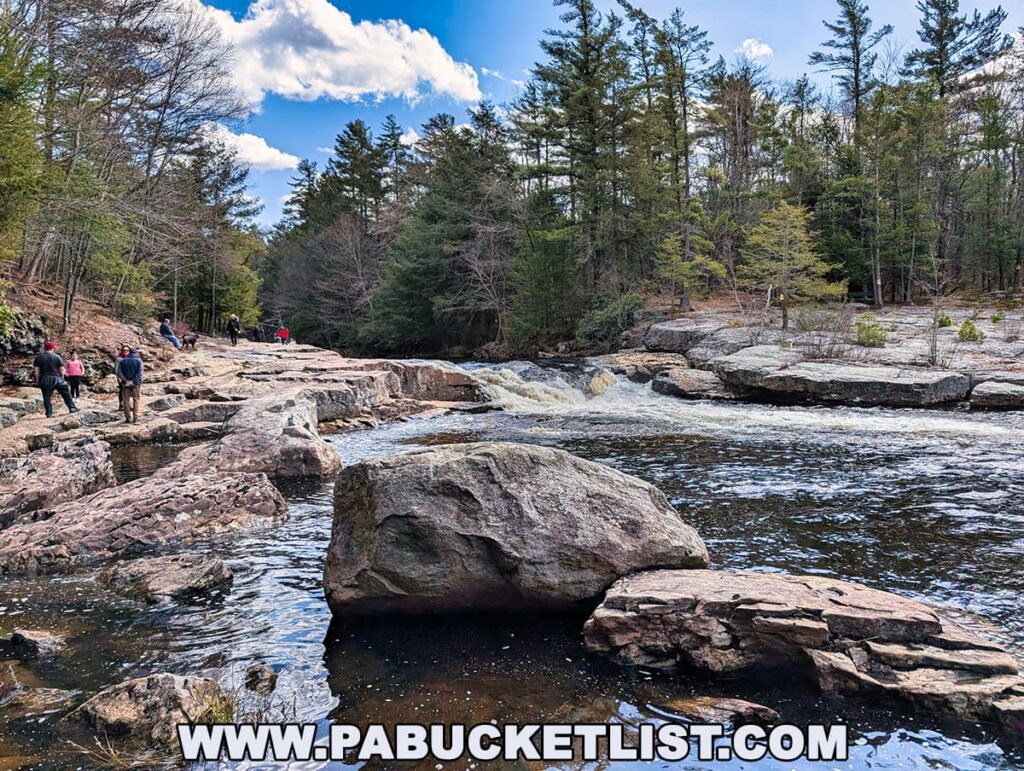 Visitors walk along large, flat rock slabs beside the rushing waters of Tobyhanna Creek at the Austin T. Blakeslee Natural Area in Monroe County, Pennsylvania, with a small waterfall visible in the distance and tall evergreens lining both sides of the creek under a blue sky.